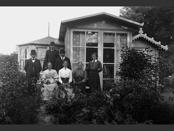 Svartvitt historiskt foto föreställande en familj i tidstypiska fina kläder framför en lummig koloniträdgård.