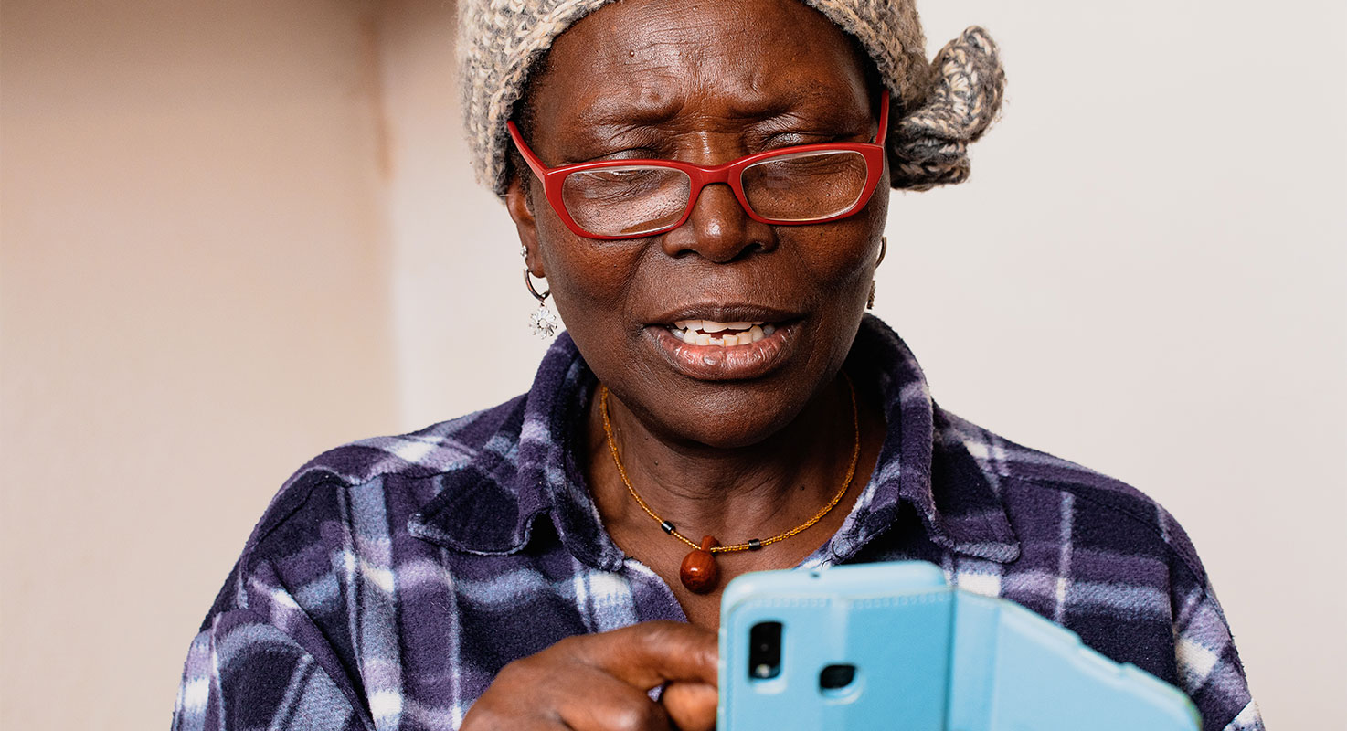 Mörkhyad kvinna över femtio med stickad mössa och röda glasögon. Kvinnan bläddrar i en mobiltelefon med ljusblått fodral.