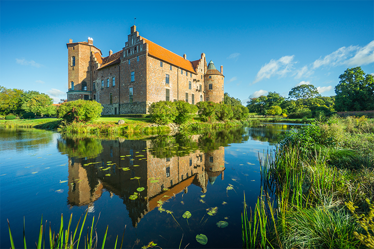 Torups slott är i mitten av bilden omringat av spegelblankt vatten och grön natur runtom en solig vårdag. 