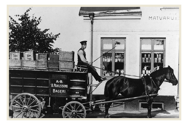 En svartvit bild av en hästdragen vagn från A.B. Pahlssons Bageri. Vagnen är lastad med korgar och lådor, och kusken sitter på sätet med tömmarna i handen. I bakgrunden syns en butik med skylten "Matvaru-", och flaskor står i fönstren.