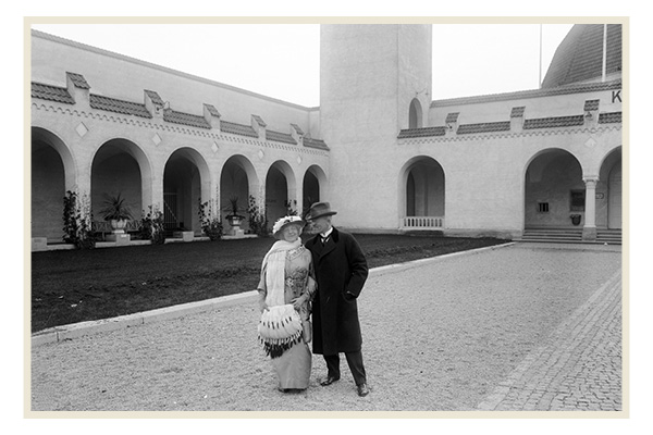 En elegant klädd man och kvinna står framför de historiska valven vid Baltiska utställningen i Malmö 1914. Miljön präglas av klassisk arkitektur och stil.