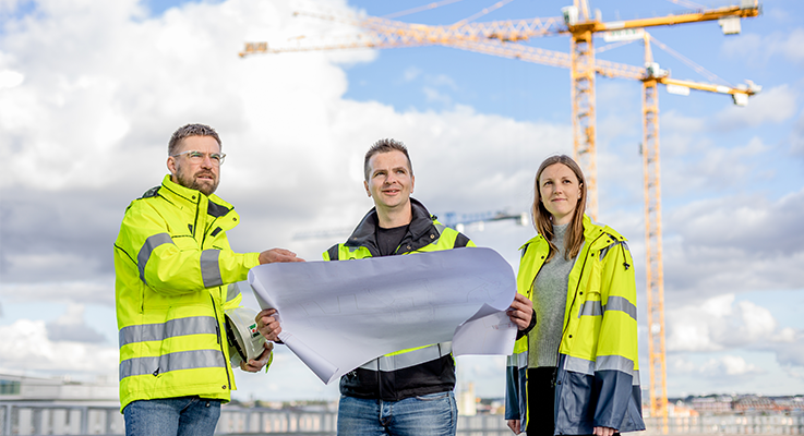 Tre medarbetare på fastighets- och gatukontoret står på en byggnad med kranar i bakgrunden. De har på sig gula arbetskläder och personen i mitten håller en ritning. 