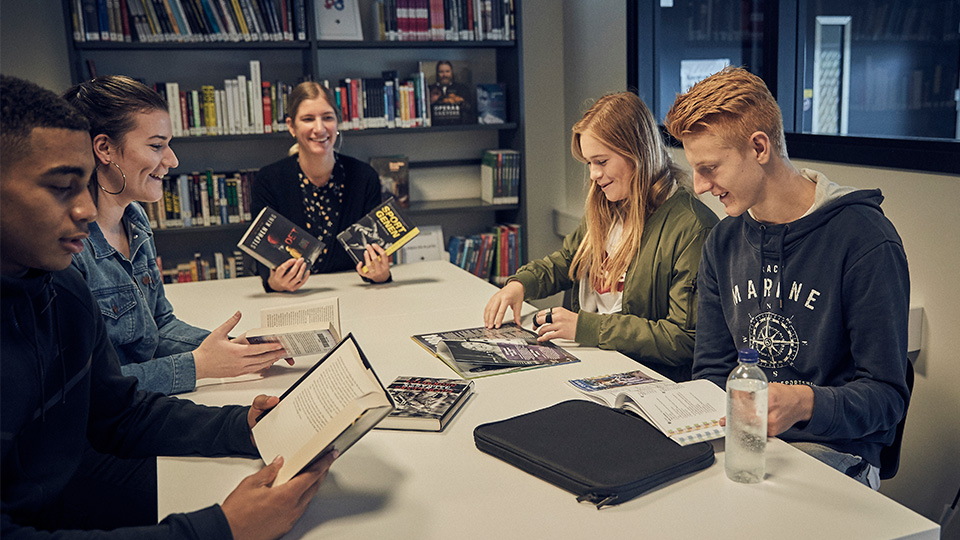 Fyra elever och en lärare sitter och arbetar i biblioteket tillsammans med en lärare.