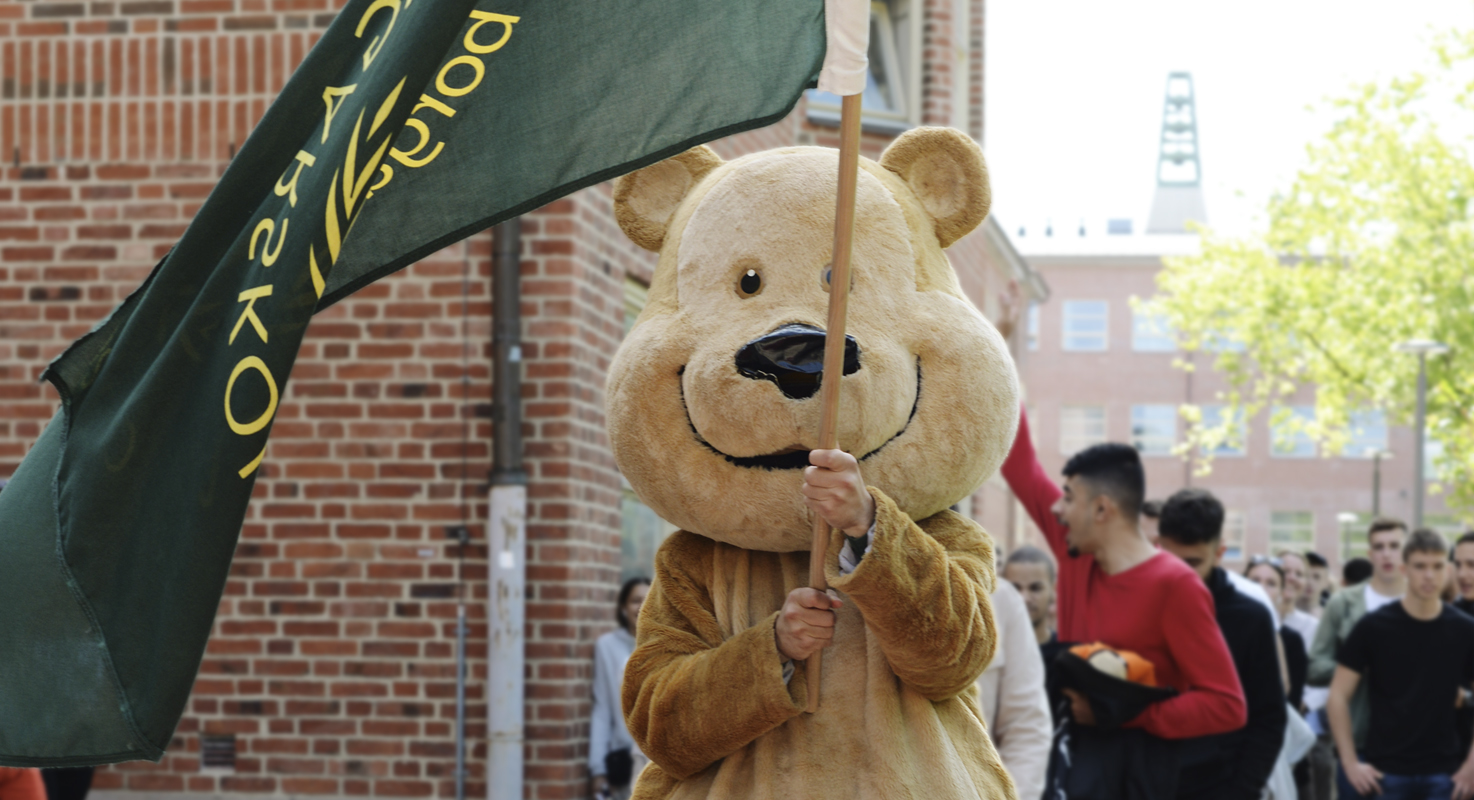 Någon iklädd en maskotdräkt som ser ut som en glad björn håller i en flaggstång med mörkgrön flagga (Malmö Borgarskolas flagga). Bakom björnen syns en stor samling elever