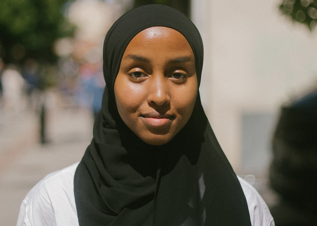 Porträttbild i färg på konstnären Ikram Abdulkadir. Hon tittar in i kameran och ler litegrann. Hon har på sig en svart slöja och en vit skjorta. Bakgrunden är oskarp men ljus med mörkare inslag.