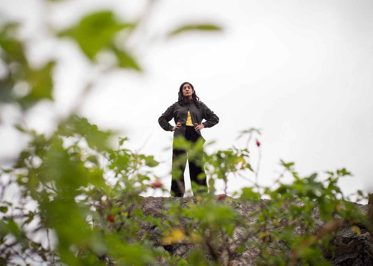 Konstnären Hanni Kamaly iklädd svarta kläder står bredbent på ett berg med armarna i sidorna. I förgrunden är det oskarpa gröna blad från en nyponbuske. Personen tittar ner in i kameran och har ett neutralt uttryck.