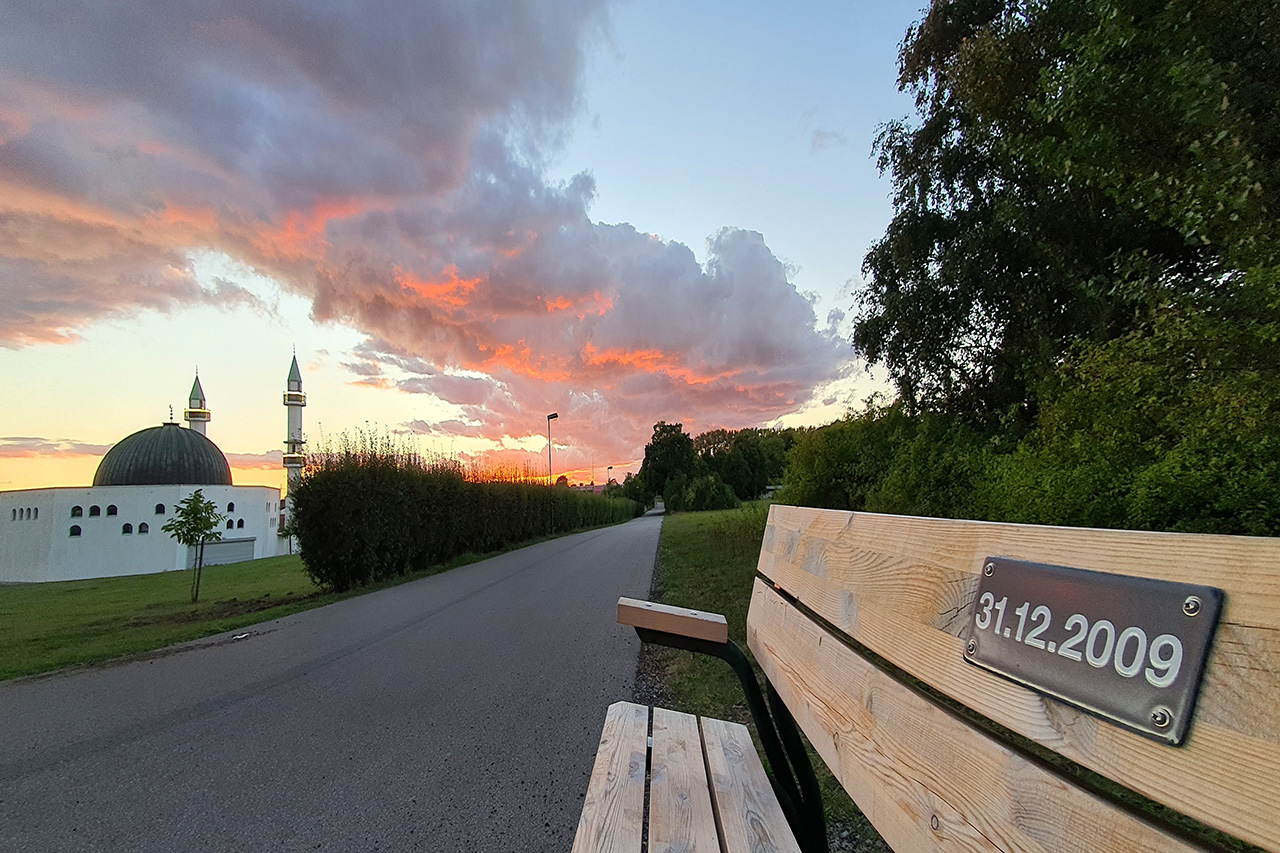 Malmö moske till vänster och en träbänk längs en gångväg med datumbeprydd plakett till höger.