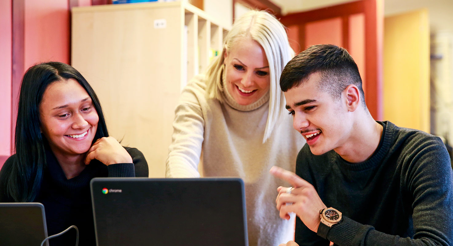 Två elever, en tjej och en kille samtalar över sina datorer. En ung lärare står mellan dem och visar på killens skärm.