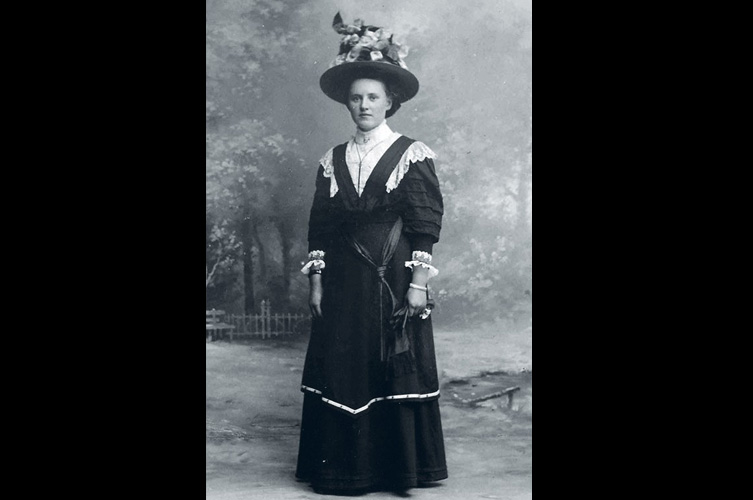 Äldre svart-vitt foto på kvinna i lång klänning och hatt. 