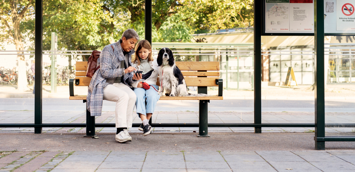 En äldre kvinna sitter vid en busshållplats i Malmö tillsammans med ett barn och en hund. 