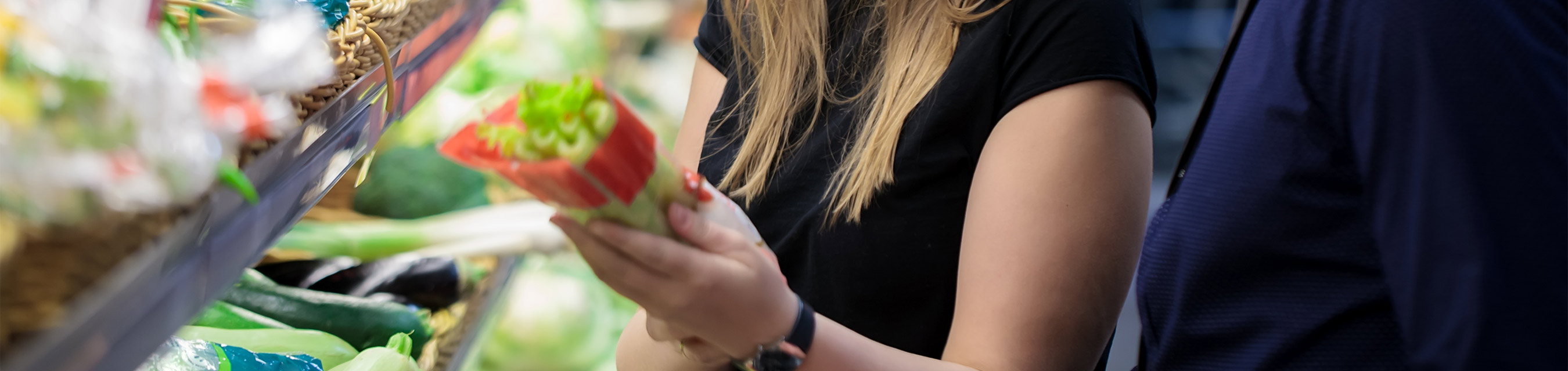 En ung tjej i svart tröja håller i ett paket selleri. Hon står framför grönsaksdisken i en butik. 