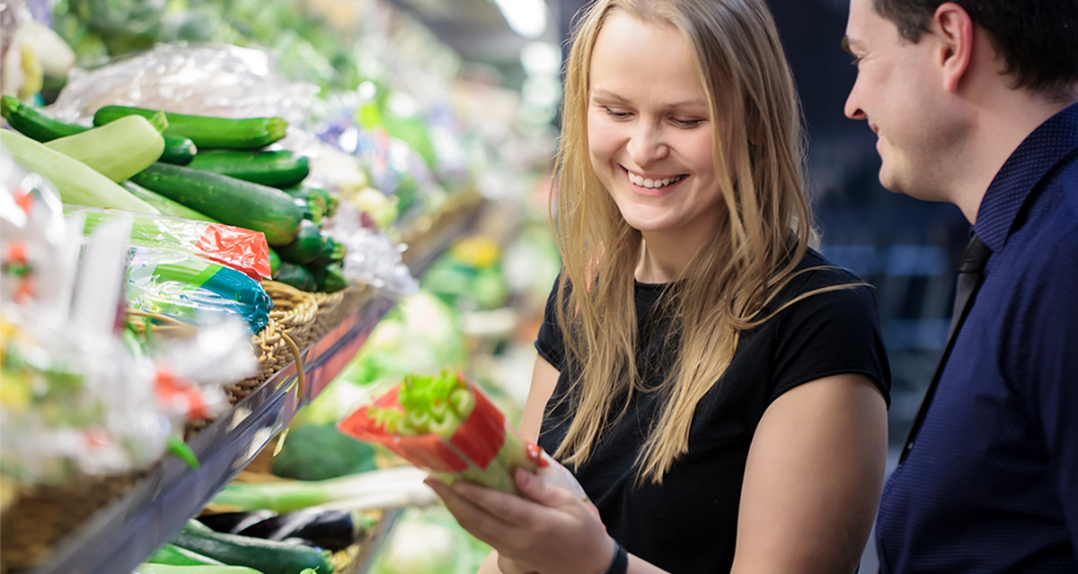 En ung tjej i svart tröja håller i ett paket selleri. Hon står framför grönsaksdisken i en butik. Snett bredvid henne står en man i blå arbetsskjorta.