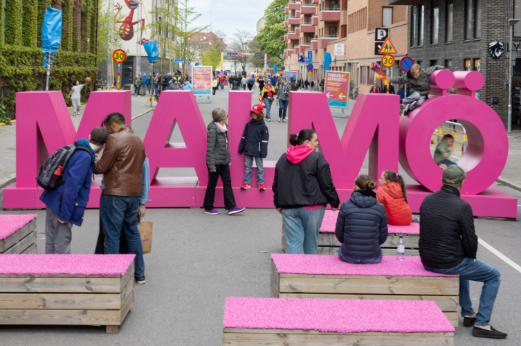 En stor rosa skylt där det står Malmö på och människor i bakgrunden som är runt skylten, sitter vid den och barn som leker mellan bokstäverna. 