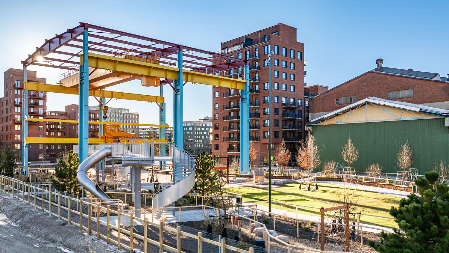 Nyanlagd stadsdelspark där en stor stålkonstruktion i olika starka färger reser sig högt över gräsmatta, buskar, klätterställningar och rör samt omgivande modern bostadsbebyggelse.