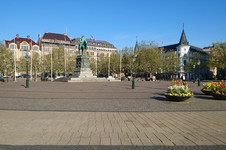 Stort öppet torg med blomsterdekorationer och flera flaggstänger, i mitten står en staty med Karl X Gustav till häst. Torget är omgivet av äldre byggnader från sekelskiftet.