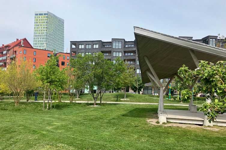 Stor gräsmatta i nyanlagd park, till höger en scenplats under tak och i bakgrunden syns de karaktäristiska byggnaderna från Hyllieområdet.