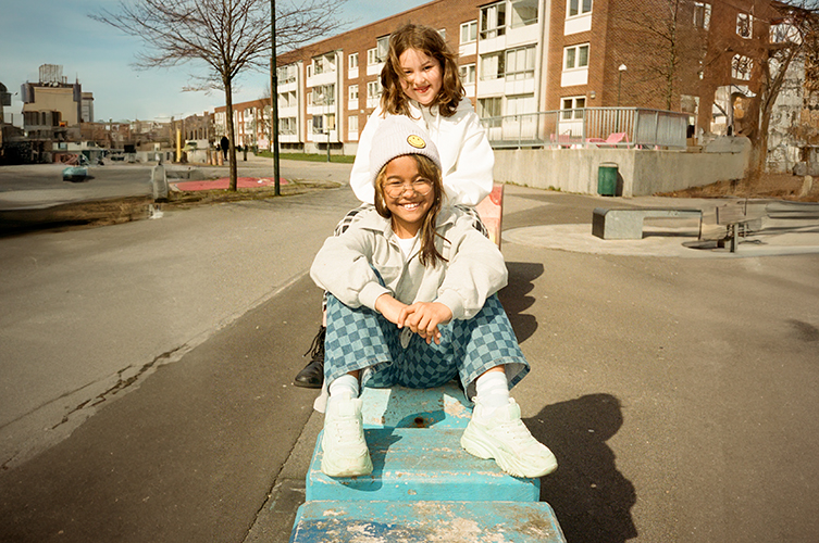 Två flickor sitter på betongfundament utomhus. Ler mot kameran. 