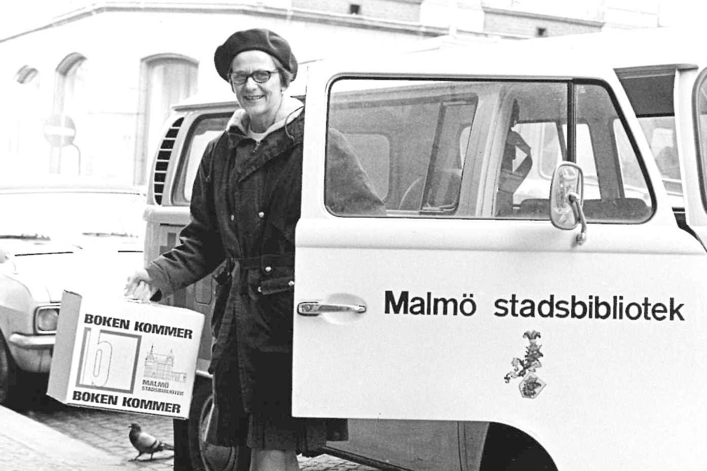 Bibliotekarie stiger ur minibuss med väska som det står "Boken kommer" på.
