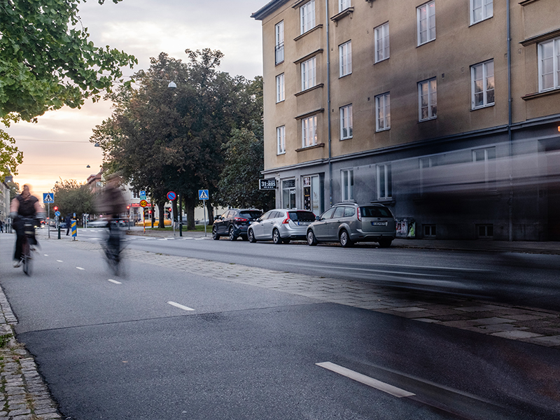 Cyklister som rör sig längs cykelbanan på Ystadsgatan, mot Möllan. I bakgrunden syns parkerade bilder. Det är fin rosa solnedgång.