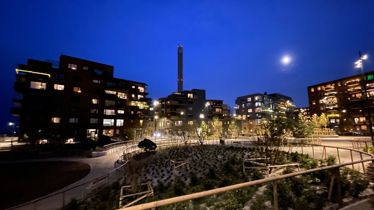 Kvällsbild från Cementkajen med mörkblå himmel och upplyst parkmiljö inramad av upplysta bostadshus.