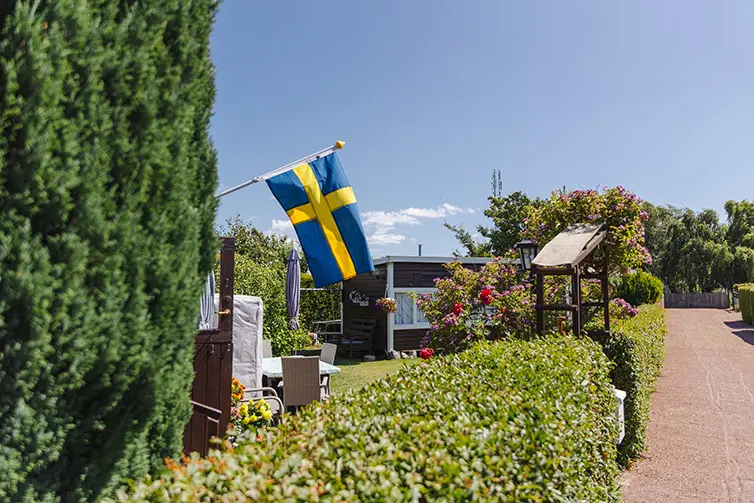 En brun kolonistuga med en trädgård på Bulltofta trädgårdsförening. I förgrunden är det ett en grind. 