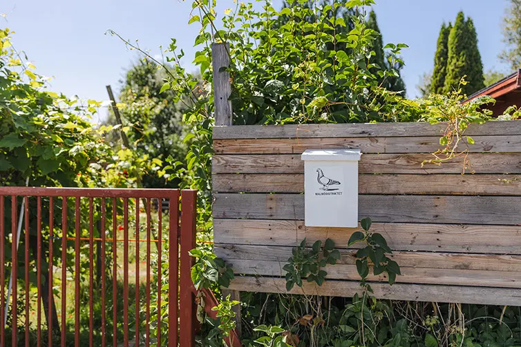 En brevlåda på ett plank bredvid en rödmålad grind. På brevlådan finns det en målad duva. 