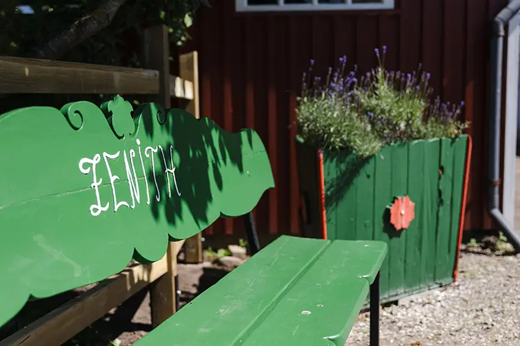 En röd stuga med en gång i mitten på Zenith koloniområde. Bredvid är det massvis med växter och grönska.
