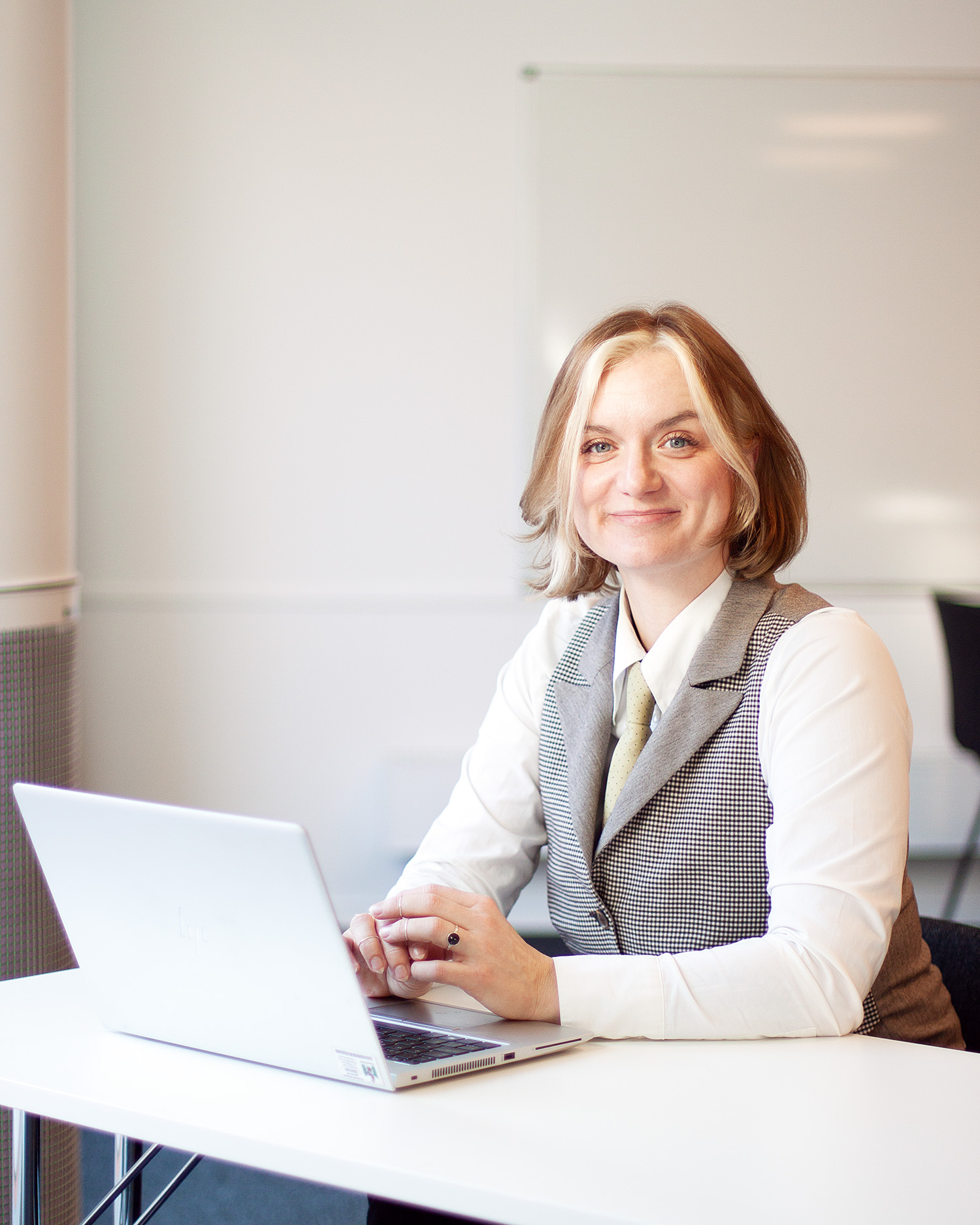 En kvinna sitter vid ett bord med en laptop. Hon tittar in i kameran och ler.