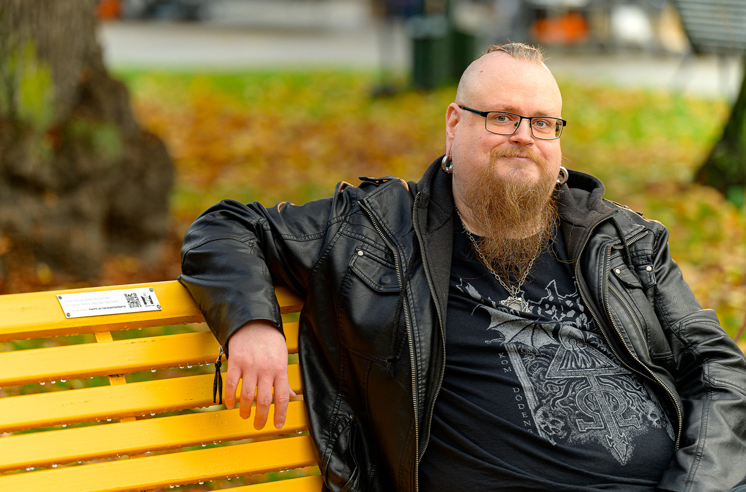 Man med skägg, glasögon, stora ringar i ögonen och svarta kläder sitter på en gul vänskapsbänk och småler mot kameran
