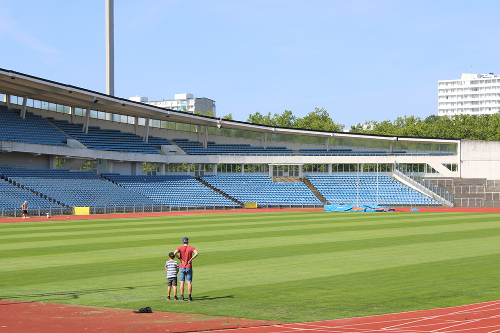 Fotografi inifrån stadion. På bilden syns men man och en yngre pojke ståendes på planen inne på stadion. I bakgrunden syns stadions blåa plaststolar.