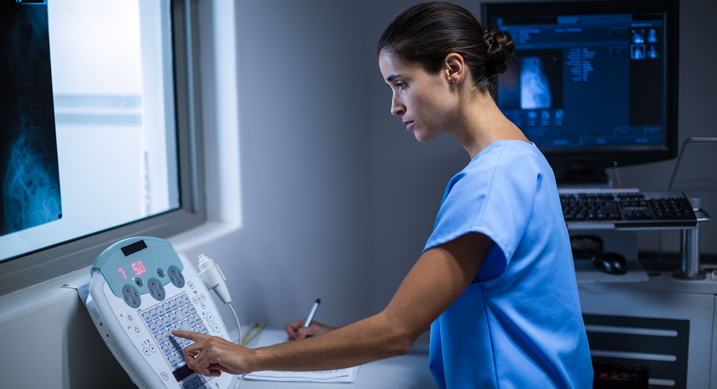 En mörkhårig kvinna i blå vårdkläder trycker på en maskin. I bakgrunden syns en mörk skärm med medicinska data.