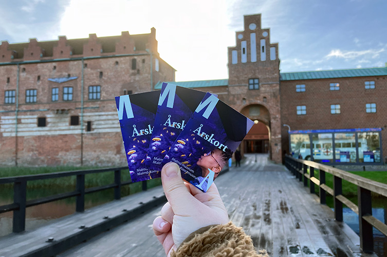 En hand som håller i tre årskort. I bakgrunden Malmöhus slott och den vita bussen.