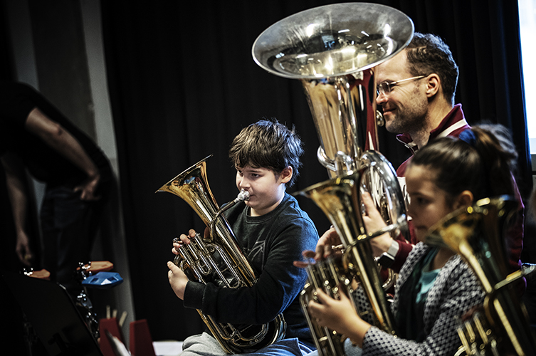 Barn och vuxna med med stora guldiga brassinstrument. De ser fokuserade ut. 