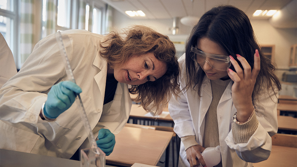 En lärare håller i en pipett och illustrerar en laboration för en elev. 