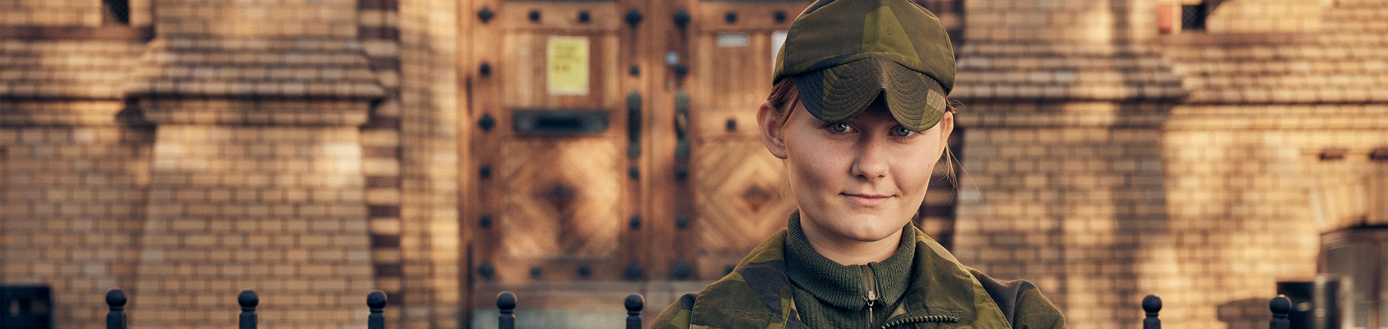 En elev i militäruniform och keps tittar leende rakt in i kameran. I bakgrunden syns skolfasad och ingång.