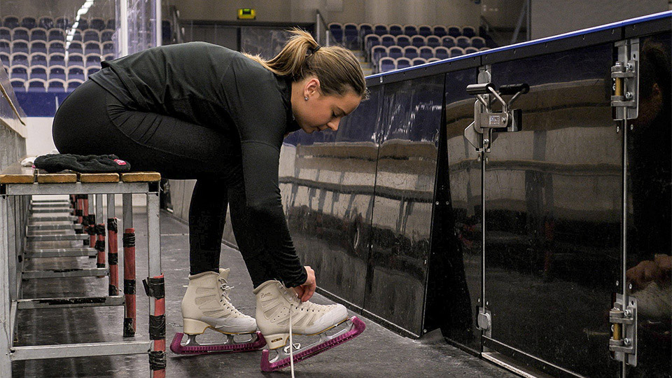 En konståkare sitter på en bänk i ishallen och knyter sina skridskor.