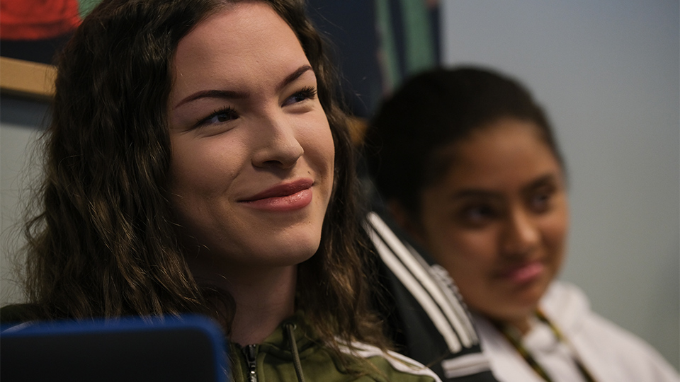 En mörkhårig tjej ser i förgrunden, med en suddig kompis i bakgrunden.