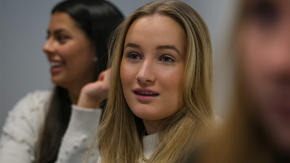 En tjej tittar framåt under en lektion.  En annan mörkhårig tjej är suddig i bakgrunden.