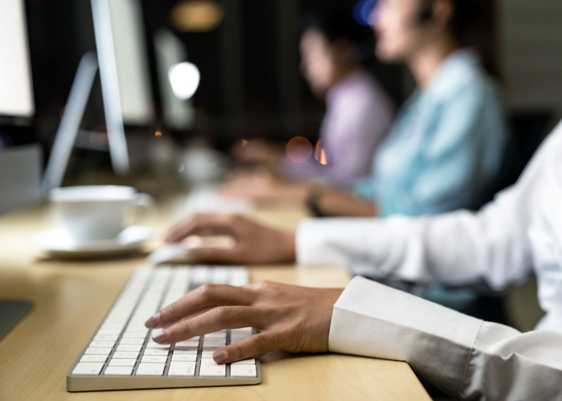 Ett callcenter med personer i rad framför datorer, i förgrunden skjortärm och händer på ett tangentbord.