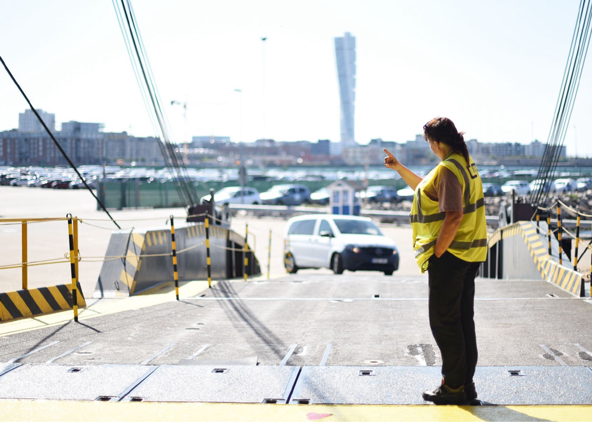 Vy över färjeramp med person iklädd reflexväst som guidar en bil på väg ombord på färjan. Del av hamnområdet, Malmö och Turning Torso i bakgrunden.