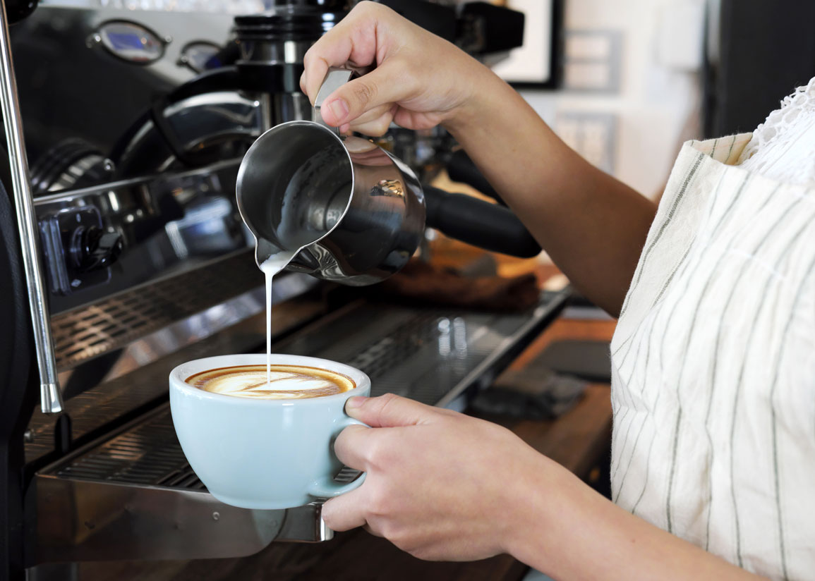 En barista häller ångad mjölk i en stor kaffekopp, framför en stor, blank espressomaskin.