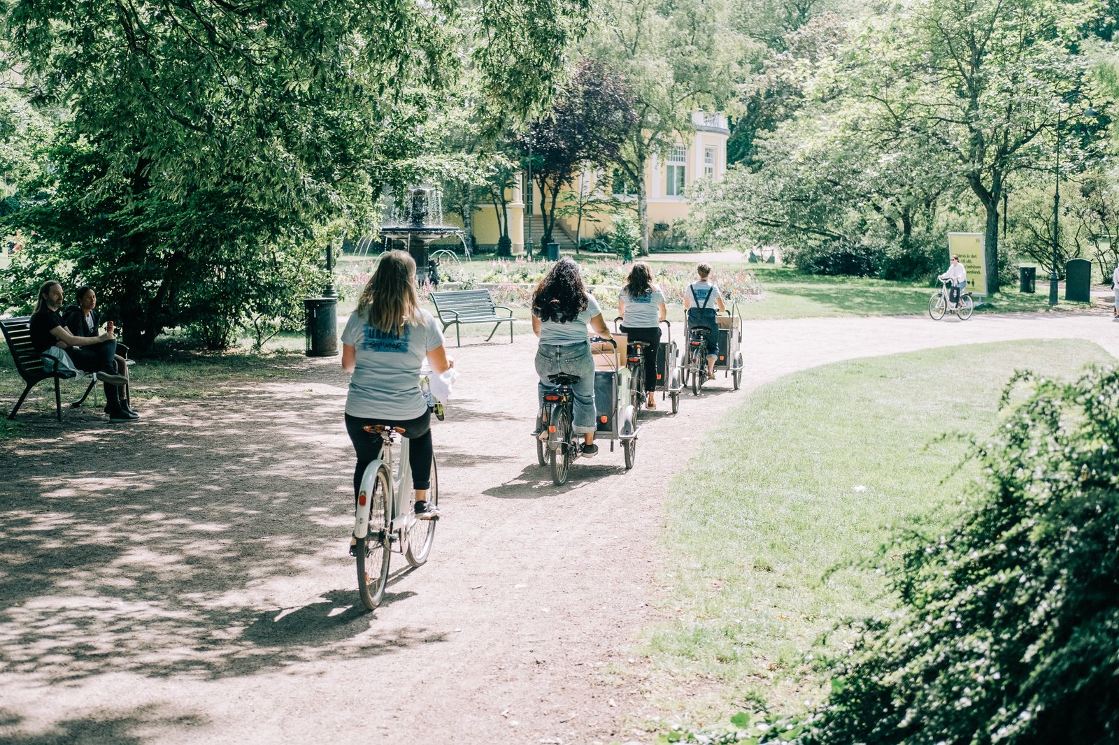 En grupp medarbetare från fastighets- och gatukontoret cyklar genom en grönskande park, varav några använder lastcyklar. I bakgrunden syns en fontän och en äldre byggnad omgiven av träd.