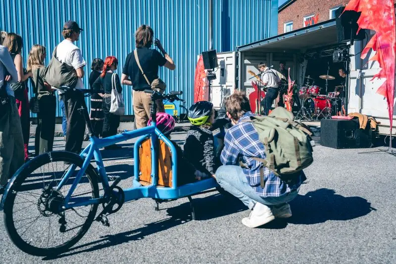 En grupp människor står och lyssnar på en livekonsert utomhus, framförd från en container. I förgrunden syns en blå lådcykel med två barn som sitter i lastutrymmet och tittar på bandet.