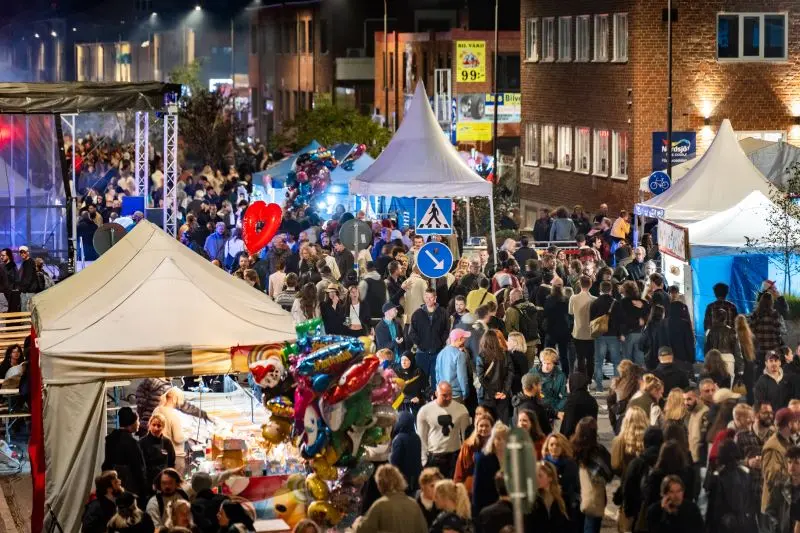 En stor folksamling på NGBG med tält och stånd längs en gata. Människor promenerar runt, medan färgglada ballonger och hjärtformade dekorationer syns i förgrunden.