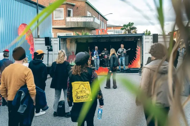 En grupp människor står och tittar på ett band som spelar musik från en scen i en container utomhus. En person bär en jacka med ett gult märke på ryggen.