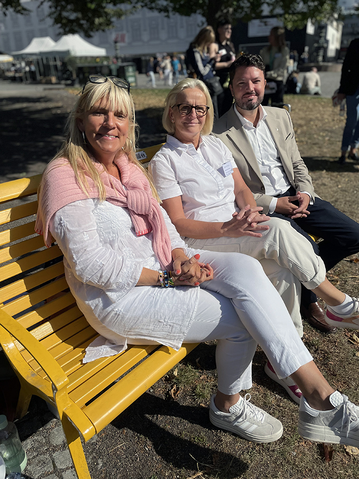 Tre personer sitter på en gul bänk och ler mot kameran