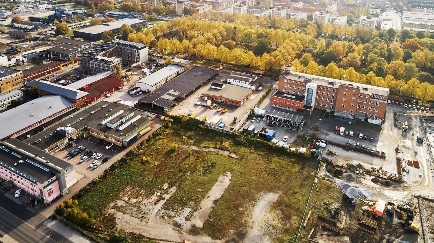 Flygfoto över ett industriområde med flera byggnader, parkeringsplatser och en stor öppen markyta i förgrunden. Träd i höstfärger bildar en gräns mellan industribyggnaderna och bostadsområdena i bakgrunden. Området verkar vara under utveckling, med pågående byggarbeten och tomma ytor för byggprojekt.