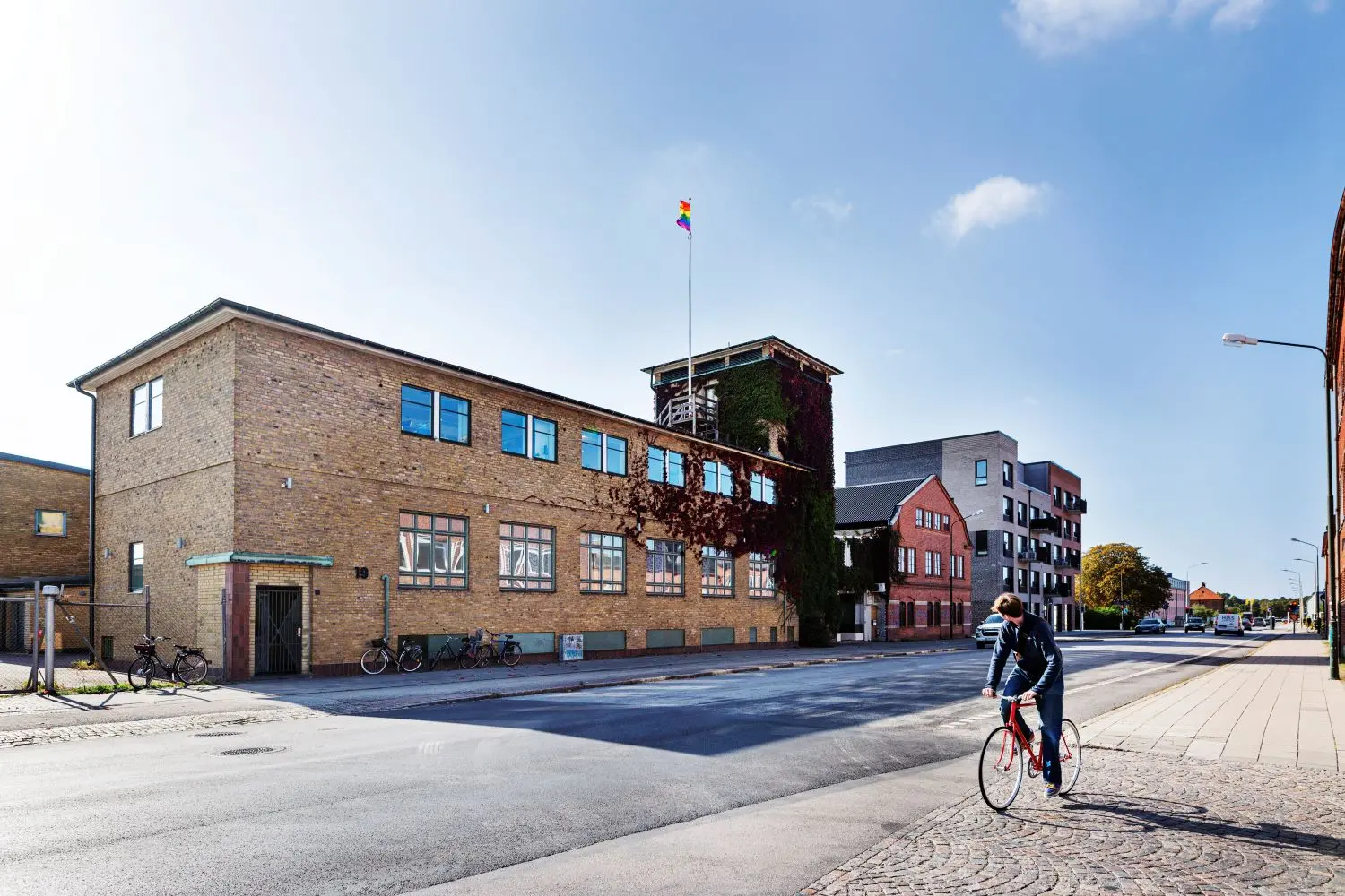 Bild av en tegelbyggnad längs en gata med en regnbågsflagga hissad på taket. Byggnaden har klätterväxter på ena sidan och flera cyklar parkerade längs trottoaren. En person på cykel rör sig längs gatan, och i bakgrunden syns fler moderna byggnader med industriell arkitektur i olika färger.