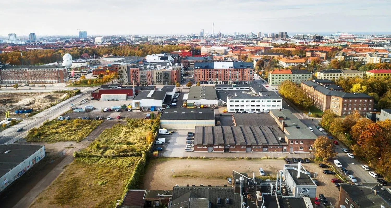 Flygfoto över ett industriområde i förgrunden, med byggnader, lagerlokaler och parkeringsplatser. I bakgrunden syns bostadskvarter med flerfamiljshus, grönskande träd och höstfärger, som skapar en kontrast till industrimarkens öppna och delvis övervuxna ytor. Längre bort sträcker sig Malmös stadssilhuett, med höga byggnader och landmärken som Turning Torso synliga vid horisonten.