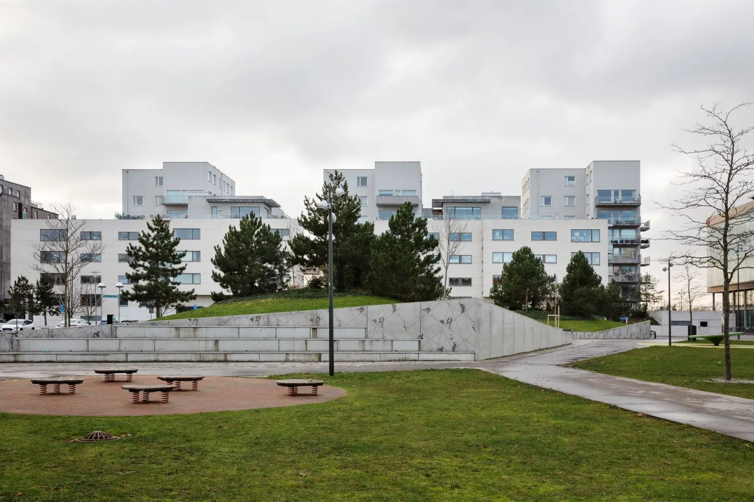 En modern bostadsbyggnad i vitt med stora fönster och balkonger omges av grönytor och träd. Framför byggnaden finns en betongmur med dekorationer och en rund yta med sittplatser. Det är en mulen dag, och gångvägar slingrar sig genom gräsmattan.